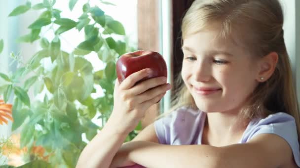 Mädchen schnüffelt am roten Apfel. Kind, das auf einem Fensterbrett am Fenster sitzt. Zoomen — Stockvideo