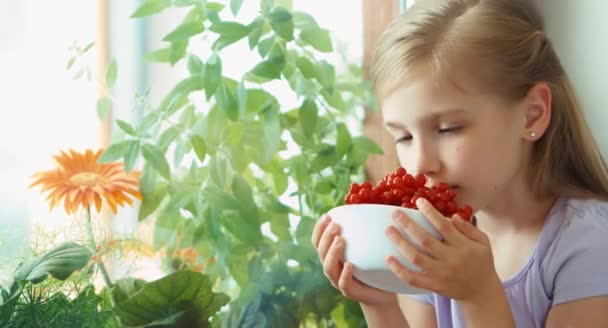 Girl showing bowl of very tasty sweet red currants and nods at camera. Child sitting on the windowsill. Closeup — Stock Video