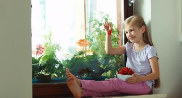 Girl sniffing very tasty sweet red currants and smiling at camera. Child sitting on the windowsill — Stockvideo