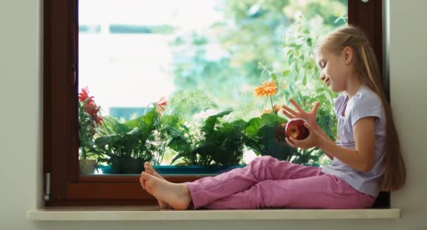 Girl playing with red apple and sniffing fruit. Child sitting on a window sill near the window — ストック動画