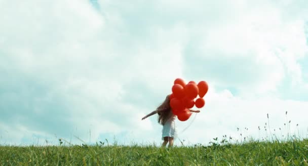 Chica en un vestido blanco sosteniendo globos rojos y girando contra el cielo en tiempo nublado — Vídeos de Stock