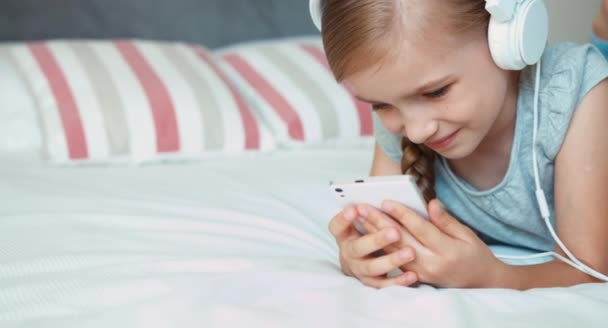 Κινηματογράφηση σε πρώτο πλάνο το παιδί πορτρέτο ξαπλωμένη σε ανάπαυση στο κρεβάτι. Κορίτσι ακούγοντας μουσική στα ακουστικά και να χρησιμοποιούν κινητό τηλέφωνο smartphone — Αρχείο Βίντεο