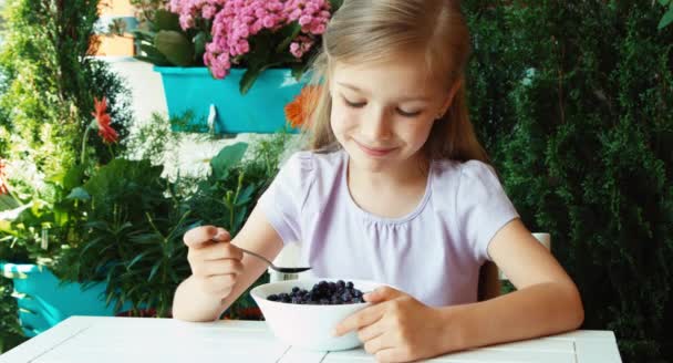 Girl eating very tasty blueberries with sugar. Child sitting resting on the table in the garden — Stockvideo