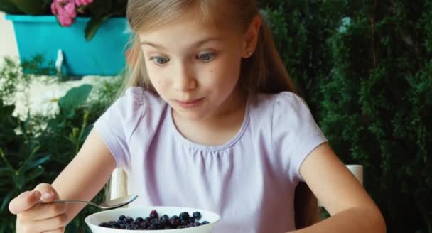 Girl eating blueberries with sugar and showing tongue. Child sitting resting on the table in the garden — Αρχείο Βίντεο