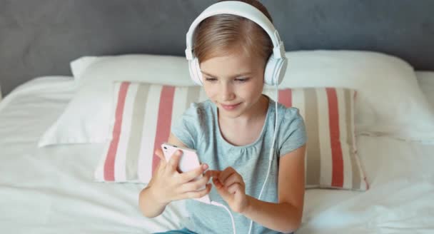 Child sitting on the bed and listening music in headphones from smartphone cell phone and dancing. Thumb up. Ok — ストック動画