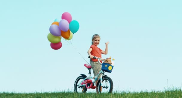 Девушка машет в камеру. Она держит велосипед с воздушными шарами. В тележке хлеб и молоко. — стоковое видео