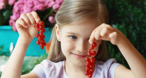 Mädchen mit einem Bund roter Johannisbeeren. Mädchen hat einen großen Teller mit roten Johannisbeeren. Kind sitzt auf dem Tisch im Garten — Stockvideo