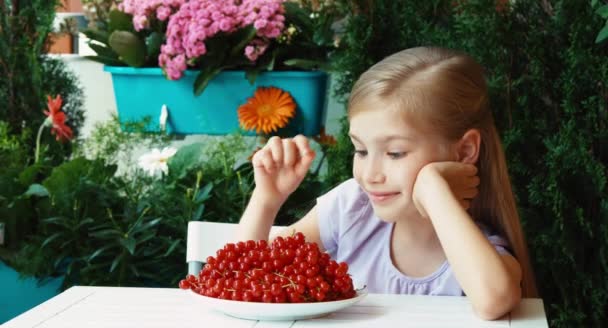 Girl showing a sprig of red currants. Girl has a big plate of red currant. Child sitting resting on the table in the garden. Thumb up. Ok — Stock Video