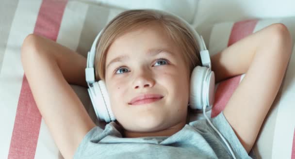 Closeup retrato menina criança ouvindo música em fones de ouvido e deitado na cama e descansando e sorrindo para a câmera — Vídeo de Stock