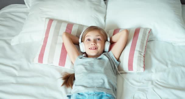 Portrait girl child listening music in headphones and lying on the bed — ストック動画