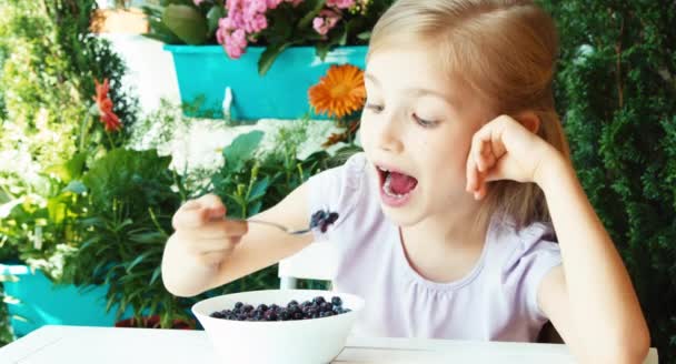 Girl eating blueberries with sugar. Child sitting resting on the table in the garden — Stockvideo