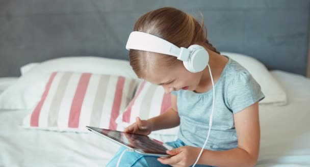 Girl using playing tablet pc and listening music in headphones. Child sitting on the bed and laughing at camera — Stockvideo