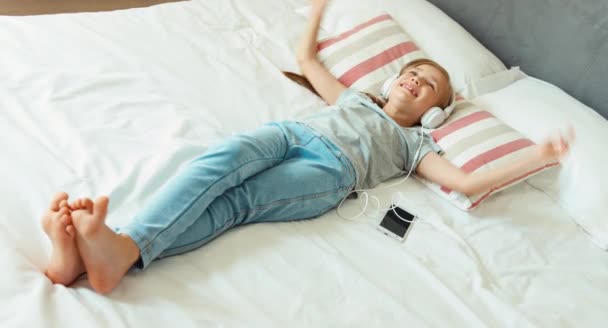 Chica escuchando música en auriculares blancos y acostada en la cama — Vídeo de stock