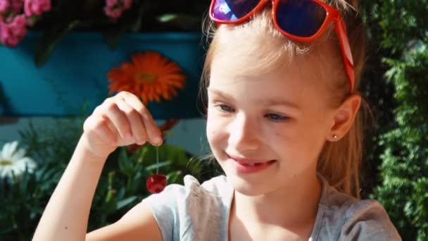 Meisje eten een kers. Kind eten bessen en bewondert hen en met de camera te kijken en te zwaaien hand — Stockvideo