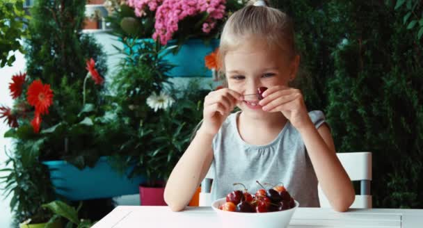 Chica comiendo cerezas rojas y sonriendo a la cámara. Grandes ojos. Cerezas muy sabrosas — Vídeos de Stock