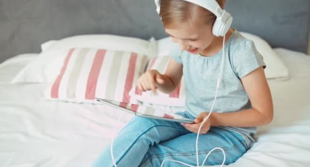 Flicka med spela TabletPC och lyssnar musik i hörlurar. Barnet sitter på sängen och ler — Stockvideo