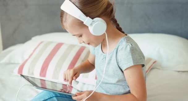 Flicka med spela TabletPC och lyssnar musik i hörlurar. Barnet sitter på sängen och skrattar — Stockvideo