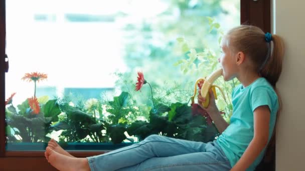 Chica comiendo plátano. Niño sentado en el alféizar de la ventana y mirando por la ventana. Ampliación — Vídeos de Stock