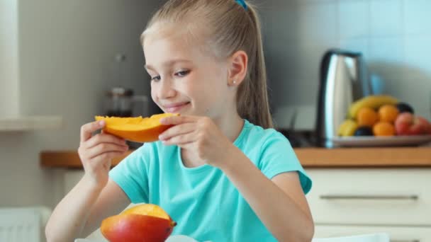 Girl sniffing and eating mango. Child sitting at the table in the kitchen and looking at camera — Stock Video