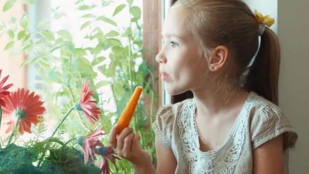 Zbliżenie portret dziewczyny jedzenie marchew i uśmiecha się do kamery. Dziecko siedząc na parapecie. Dziecko, które kocha zdrowej żywności. Powiększanie — Wideo stockowe