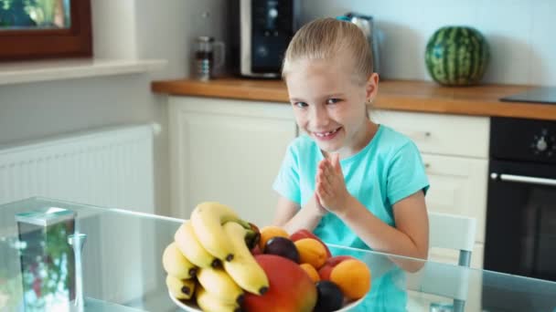 Ragazza promozione frutta. La ragazza gli strofina le mani. Ragazza con un grande piatto di frutta seduta al tavolo della cucina. Guardando la macchina fotografica e sorridendo. Zoomare — Video Stock