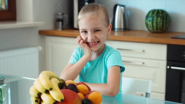 La ragazza fa pubblicità alla frutta. Bambino seduto sul tavolo della cucina e ridendo della macchina fotografica. Pollice in alto. Va bene. Zoomare — Video Stock