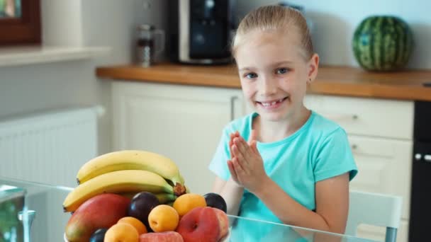 Girl rubs his hands. Girl with a big plate of fruits sitting at the kitchen table. Looking at camera and smiling. Zooming — Stock Video