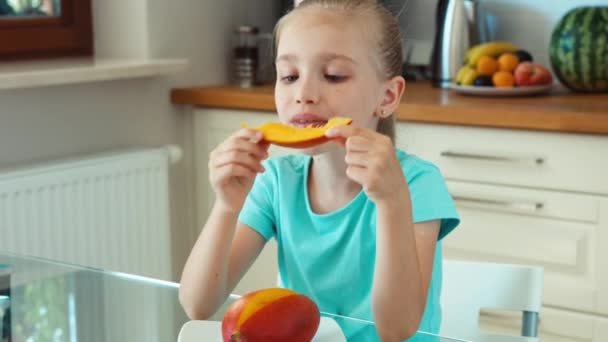 Chica comiendo mango y sonriendo a la cámara. Niño sentado a la mesa en la cocina. Ampliación — Vídeos de Stock