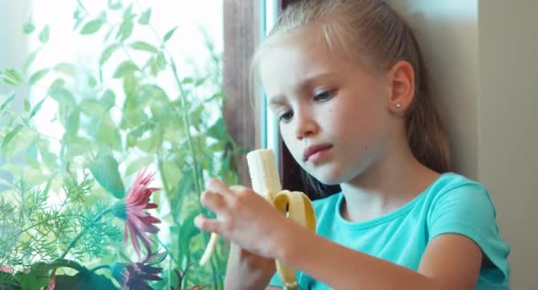 Menina retrato close-up comendo banana e sentado no peitoril da janela e olhando pela janela e olhando para a câmera. Polegar para cima. Está bem. — Vídeo de Stock