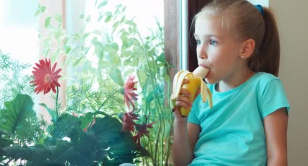 Πορτρέτο κορίτσι τρώει μπανάνα και κάθεται στο περβάζι και κοιτάζοντας έξω από το παράθυρο και βλέπει τα φωτογραφικών μηχανών. Τον αντίχειρα επάνω. Ok — Αρχείο Βίντεο