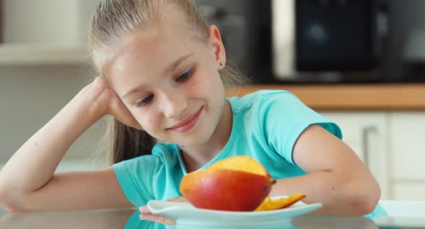 Portrait girl looking at mango and camera. Child promotes fruit mango. Child sitting at the kitchen table — Stock Video