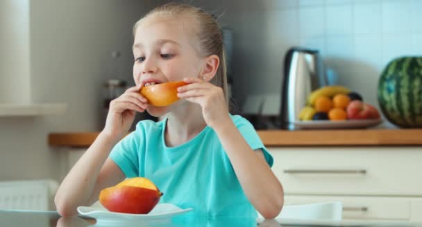 Girl eating mango. Child sitting at the table in the kitchen and looking at camera. Thumb up. Ok — Stock Video