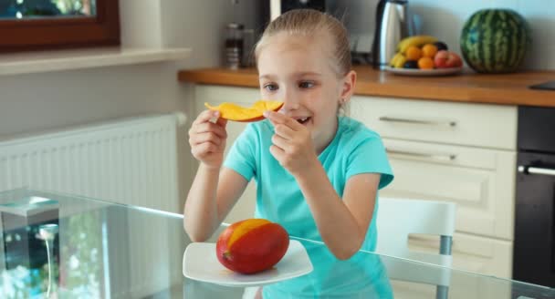 Meisje eten mango en glimlachend in de camera. Kind op de tafel in de keuken — Stockvideo