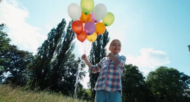 Girl touching balloon and smiling at camera against the sun. Child with balloons in the park — Stock Video