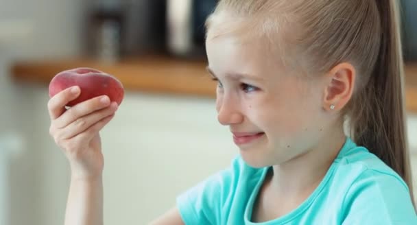 Nahaufnahme Porträt Mädchen schnüffelt Pfirsich. Kind sitzt am Küchentisch und lächelt in die Kamera — Stockvideo