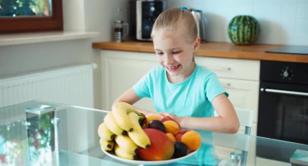 Rapariga anuncia fruta. A rapariga esfrega-lhe as mãos. Menina com um grande prato de frutas sentado na mesa da cozinha. Olhando para a câmera e sorrindo — Vídeo de Stock