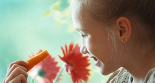 Extremo primer plano retrato chica comiendo zanahoria y sonriendo a la cámara. Niño sentado en un alféizar de ventana. Niño que ama la comida saludable. Niño mira la zanahoria — Vídeos de Stock