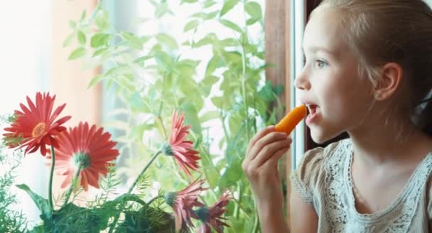 Detailní portrét dívka jíst mrkev a usmívá se na kameru. Palec nahoru. Ok. Dítě sedí na okenním parapetu — Stock video