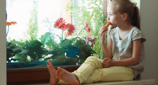 Tjej äter morot och ler mot kameran. Barnet sitter på en fönsterbräda. Tummen upp. Okej — Stockvideo