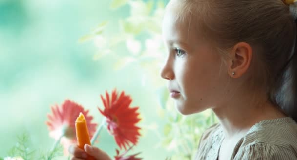 Closeup πορτρέτο κορίτσι τρώει καρότο και βλέπουν τα φωτογραφικών μηχανών. Παιδί που κάθεται σε ένα windowsill — Αρχείο Βίντεο