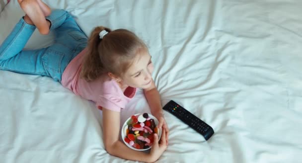 Niña solitaria cambiando la televisión acostada en la cama. Un niño sorprendió a la televisión. Un niño comiendo dulces. Vista superior — Vídeos de Stock