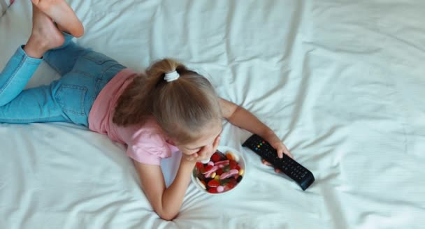 Ragazza che guarda la TV sdraiata sul letto. Un bambino che mangia dolci. Vista dall'alto — Video Stock