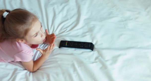 Mädchen vor dem Fernseher auf dem Bett liegend. Kind schockierte das Fernsehen und lächelte in die Kamera. Ansicht von oben — Stockvideo