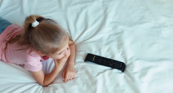 Menina assistindo TV deitada na cama. Criança chocado TV e rindo da câmera. Vista superior — Vídeo de Stock