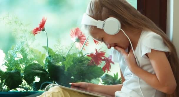 Kız tablet PC'yi kullanma ve kameraya gülme. Çocuk kulaklık müzik dinleme. Bir pencere pervazına çiçek karşı oturan çocuk — Stok video