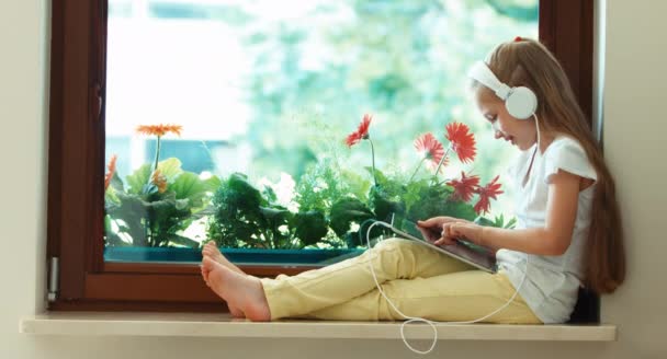 Дівчина використовує планшетний ПК. Дитина сидить на підвіконні проти квітки. Дитина слухає музику на навушниках — стокове відео