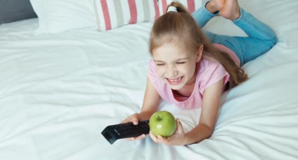 Dziewczyna, oglądać Tv leżąc na łóżku. Dziecko jedzenia apple — Wideo stockowe