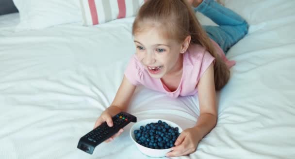 Mädchen vor dem Fernseher auf dem Bett liegend. Kind isst Blaubeeren — Stockvideo