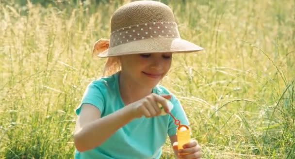 Närbild porträtt flicka blåser såpbubblor i gräset och ler mot kameran. Fontänen av bubblor — Stockvideo