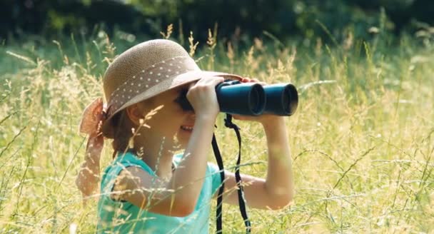 Портрет смеющегося юного натуралиста через бинокль, смотрящего дикую природу. Ребенок сидит на высокой траве — стоковое видео